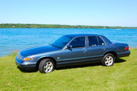 1985 Mercury Grand Marquis - A blue Mercury Grand Marquis sits on a grassy bank next to a bright blue lake under a clear sky.
