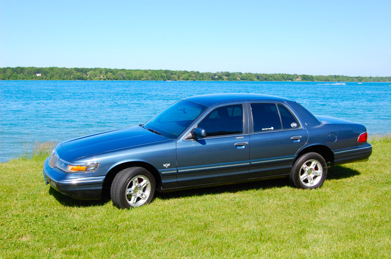 Blue Mercury Grand Marquis parked on grass by a lake. It has a sticker on the rear door