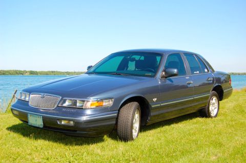 1985 Mercury Grand Marquis - A dark blue Mercury Grand Marquis sedan is parked on a grassy area next to a body of water under a clear blue sky.