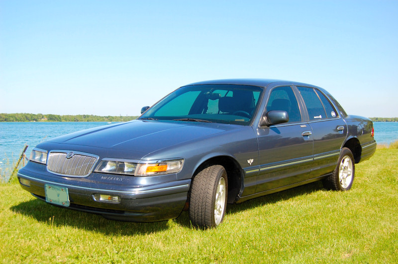 Dark blue Mercury Grand Marquis sedan parked on grass by a lake, front 3/4 view