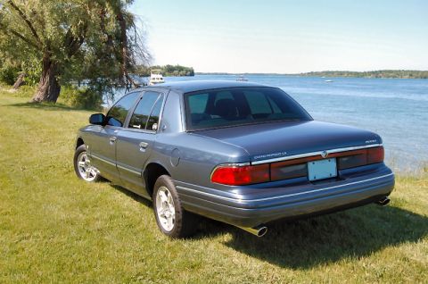1985 Mercury Grand Marquis - Rear view of a blue sedan parked on a grassy area next to a body of water.