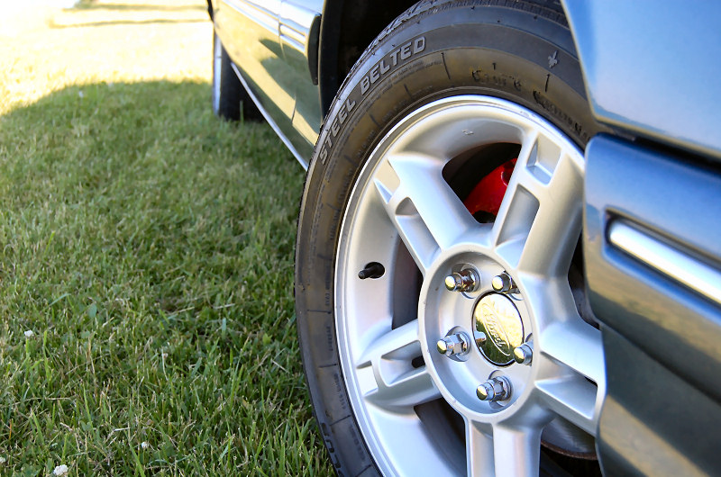 Close-up of a car's silver alloy wheel with red brake caliper visible behind it. Grass is in the background