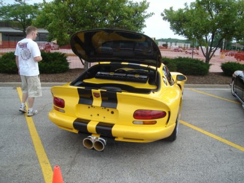 1992 Ford Crown Victoria - A yellow sports car with dual exhaust pipes and black racing stripes is parked with its trunk open, revealing audio equipment inside.