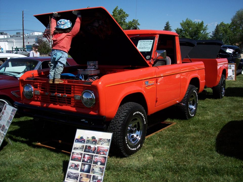 Orange 1972 Ford Bronco with hood open, child looking at engine. Another Bronco is visible behind it