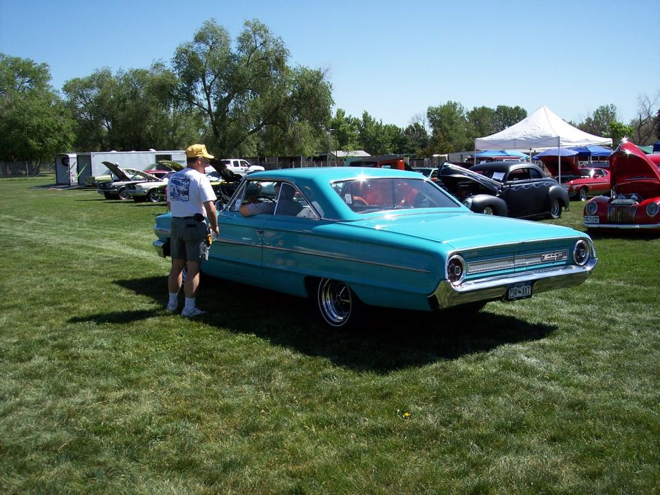 Turquoise 1964 Ford Galaxie 500 coupe parked on grass, man standing by door. Other cars and tents in background