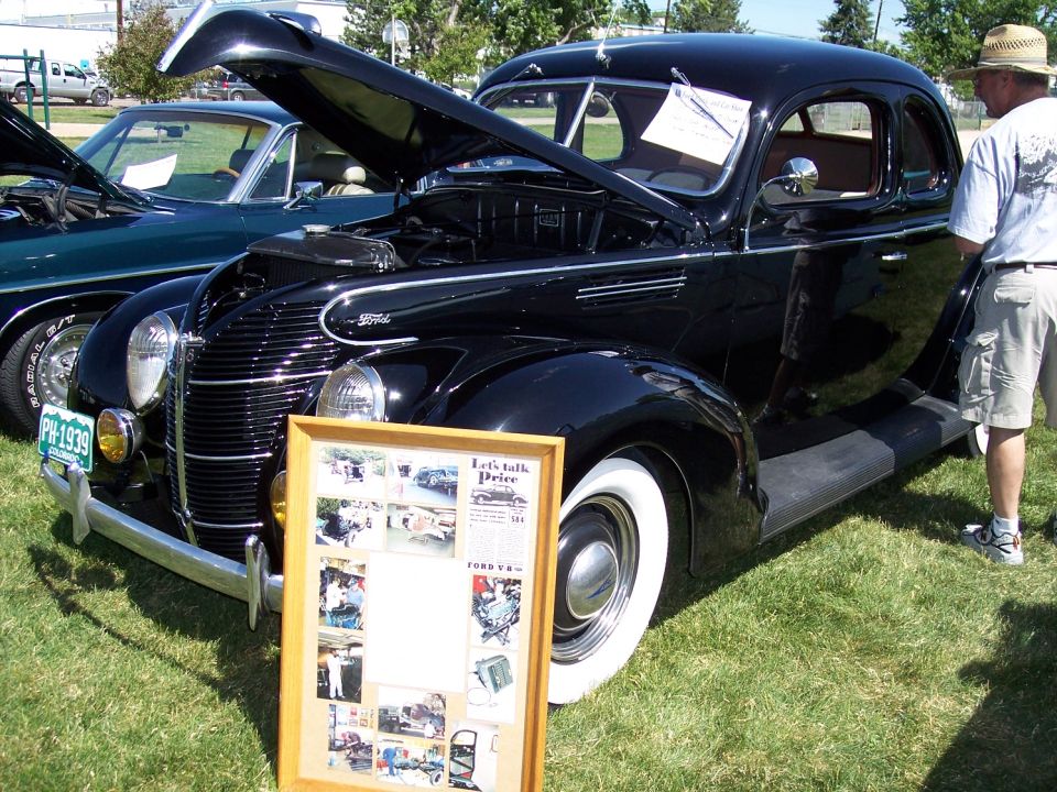 Black 1939 Ford Coupe with hood open, next to a dark green car. Display board in front of the coupe