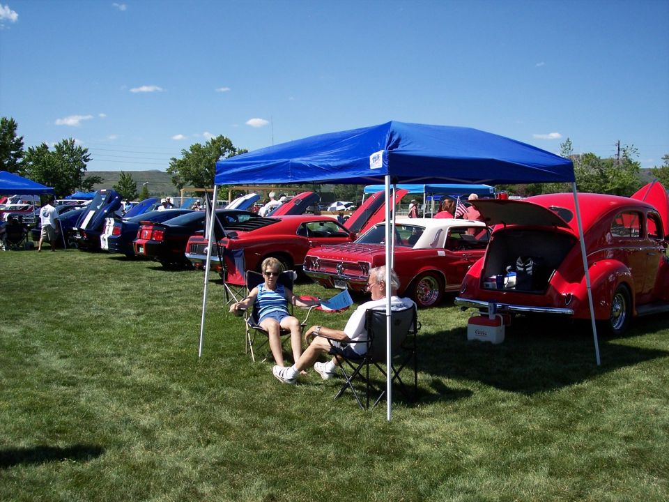 Several classic cars are parked on a grassy field under a blue canopy, with people relaxing in chairs