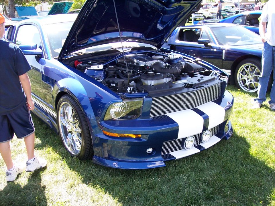 Blue Ford Mustang with racing stripes and hood open, showing engine. Another blue car is parked behind it