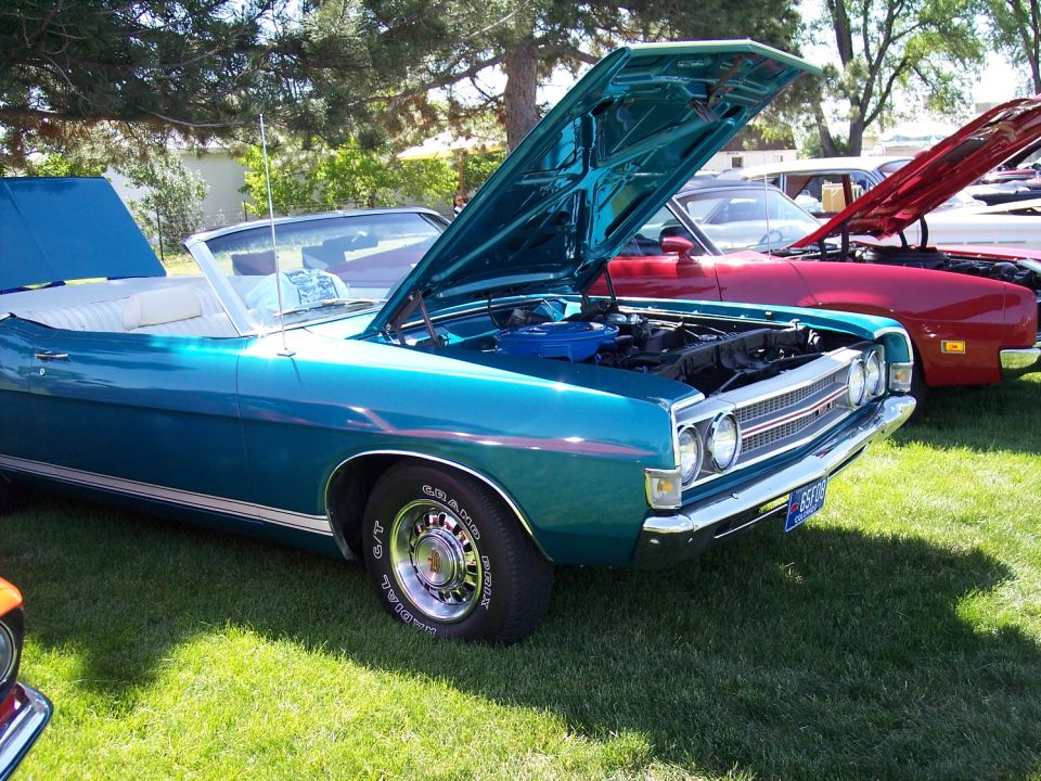 Teal 1970 Ford Torino GT convertible with hood open, parked on grass next to a red car