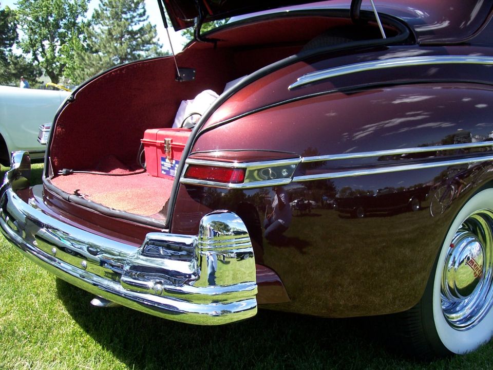 Rear view of a maroon 1940s car with open trunk showing red carpet and a red toolbox