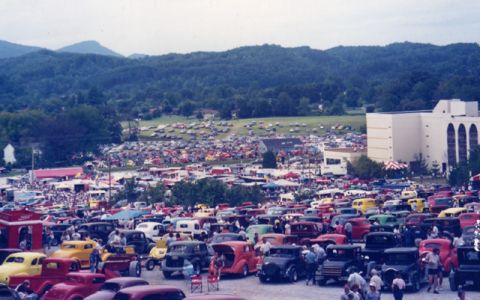1981 Ford Bronco - A large gathering of vehicles is visible in an outdoor setting, with hills and a building in the distance.