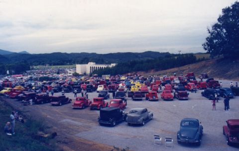1981 Ford Bronco - A large gathering of classic cars are parked in an outdoor lot, with a building and hills visible in the distance.