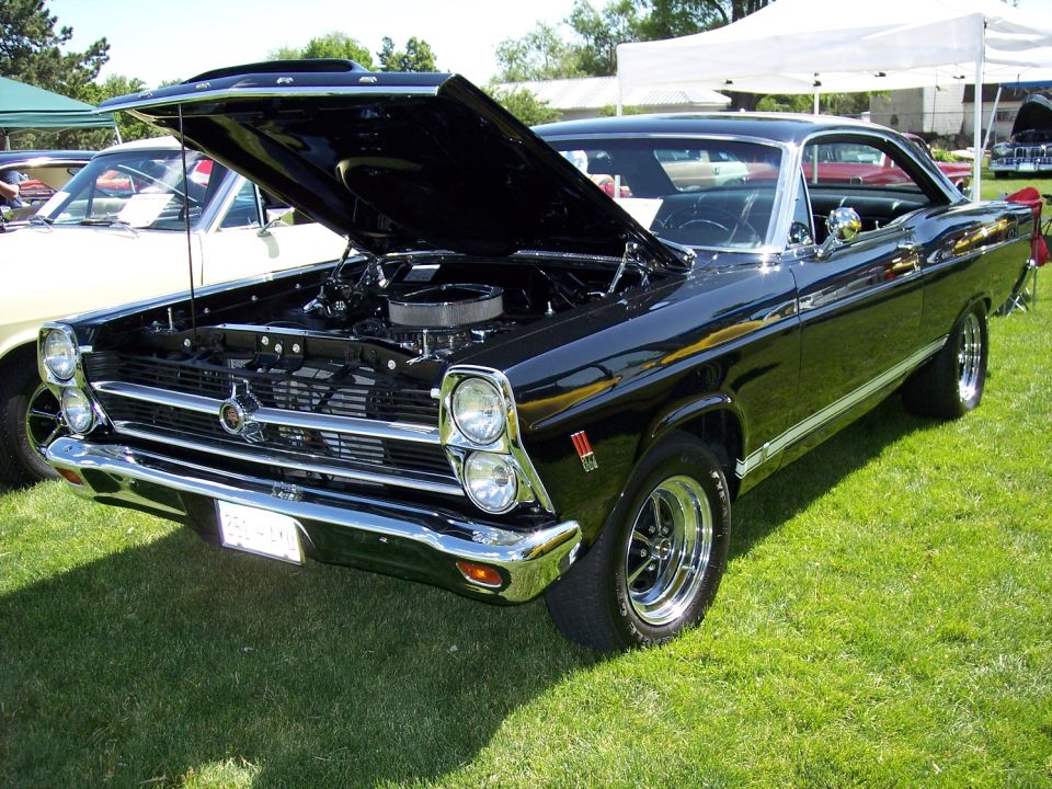 Black classic car, hood open, showing engine. Another car is parked nearby. Tents are set up in the background