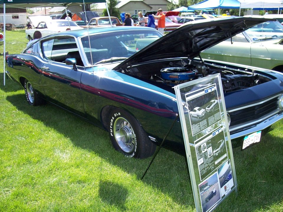 Dark blue 1970 Ford Torino GT with hood open, parked on grass at a car show. Display board in front