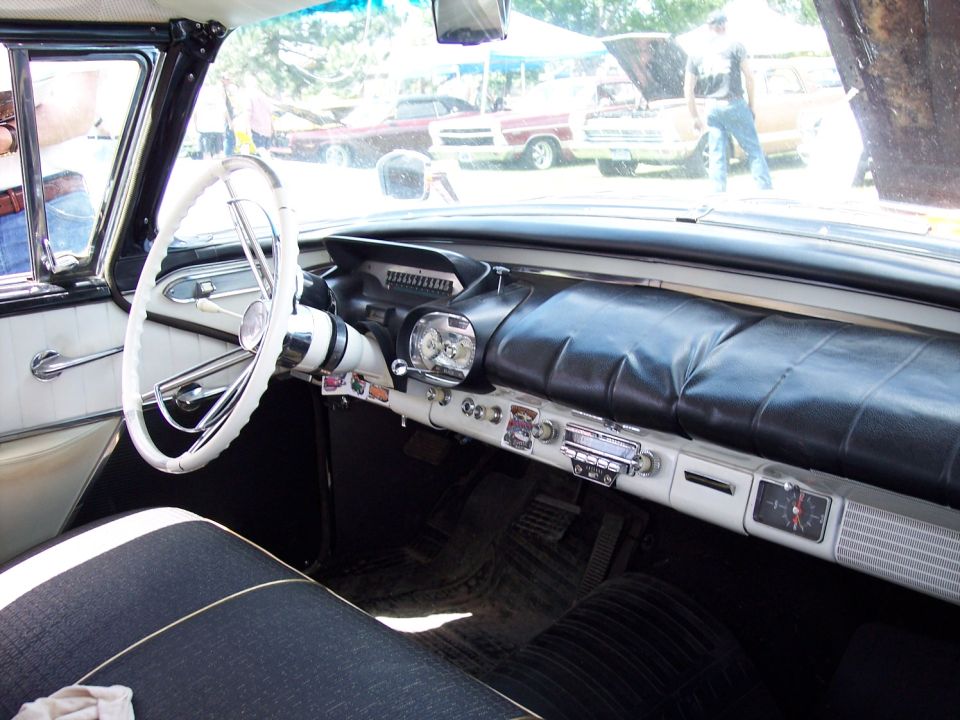 Interior view of a classic car with a white steering wheel and dashboard. Other cars visible outside