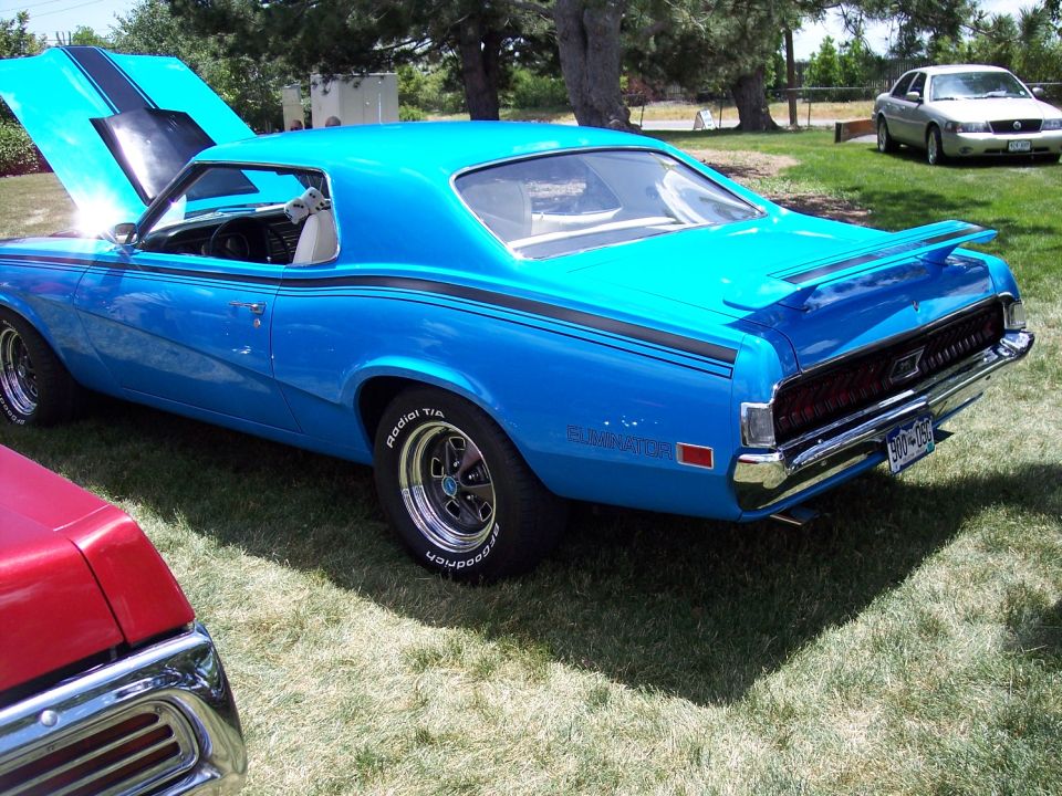 Blue 1970 Mercury Cougar Eliminator with black racing stripes, hood open. Another car is partially visible in the foreground