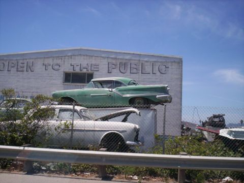 1989 Ford Bronco - Two stacked 1950s American finned sedans in a salvage yard, a weathered green two-tone hardtop perched on top of a white one, seen from the…