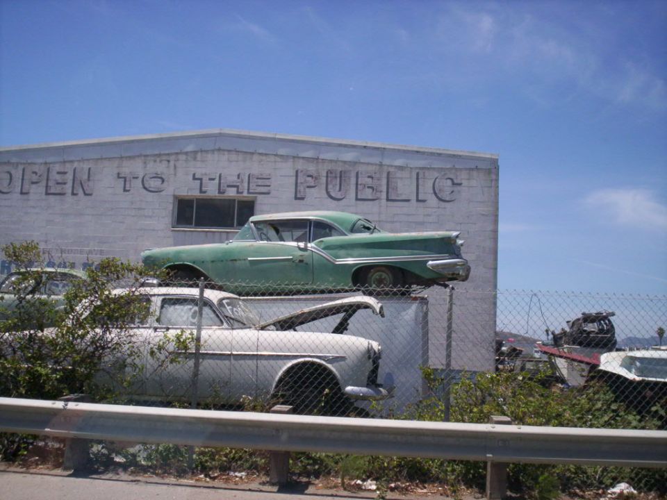 Green 1950s finned sedan stacked on a white one in a salvage yard behind chain-link fence