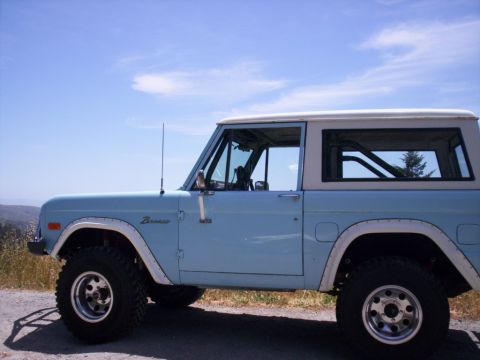 1989 Ford Bronco - Driver-side profile of a light blue early Ford Bronco with a white removable hardtop, white fender flares and rocker trim, parked on a dirt…