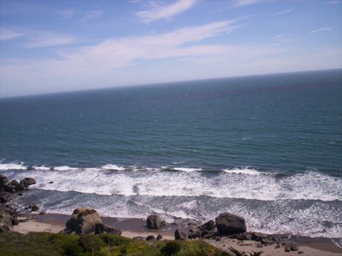 1989 Ford Bronco - Coastal overlook of a rocky beach with surf breaking on sand and dark boulders, open ocean stretching to the horizon under a partly cloudy…