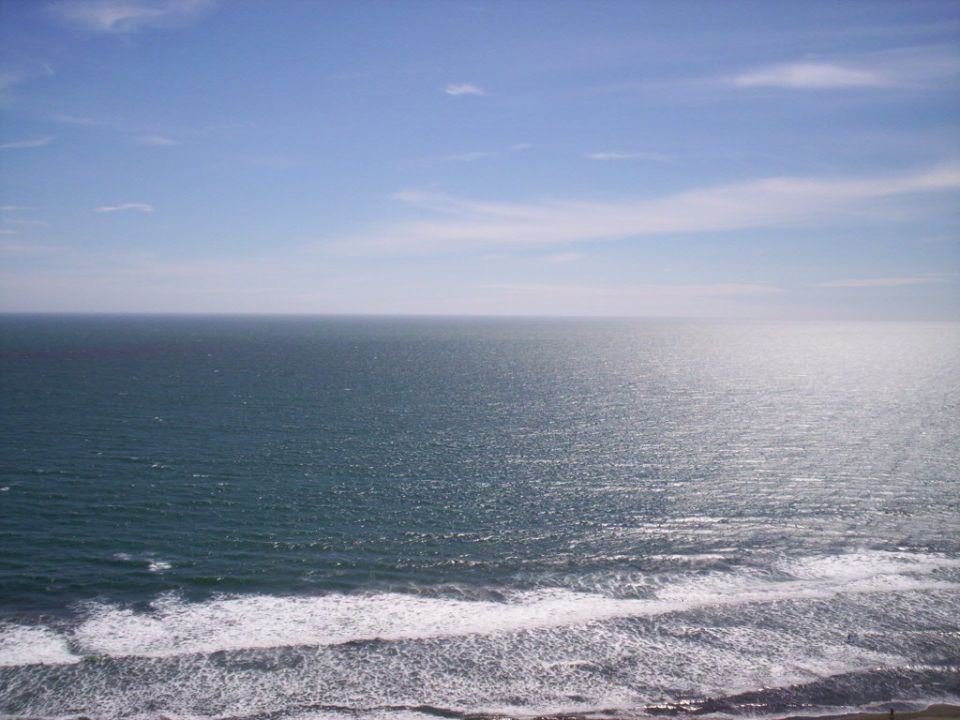 Sunlit open ocean with white surf breaking on the shore under a hazy blue sky