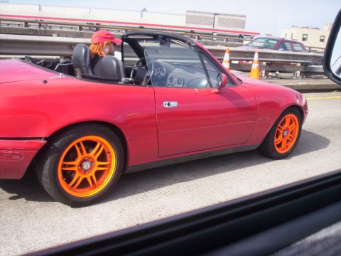 1989 Ford Bronco - A red convertible with bright orange wheels drives on a highway with another car in the background.