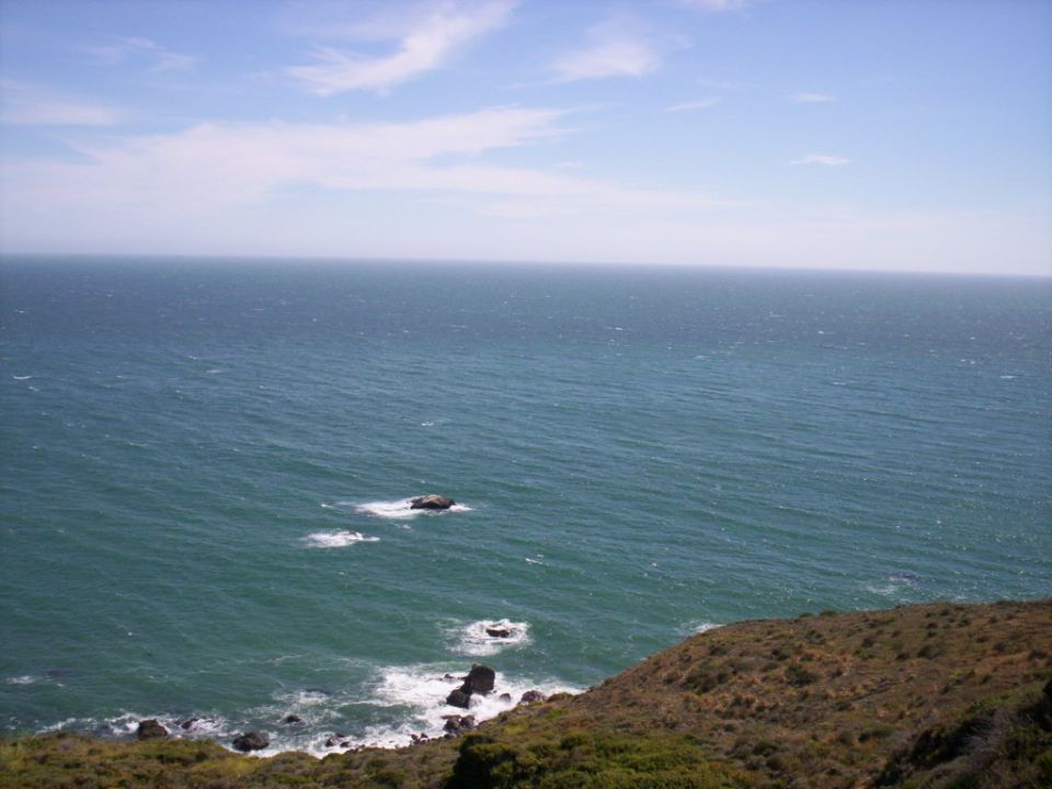 Ocean waves crash on a rocky, brush-covered coastline under a hazy blue sky