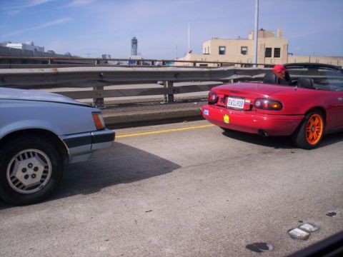 1989 Ford Bronco - Two cars driving on a highway, one is a red convertible with custom wheels and plate.