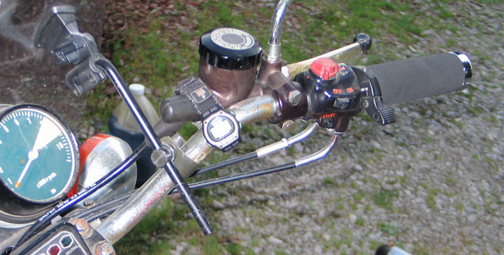 Motorcycle handlebars with a speedometer, brake reservoir, and controls. A watch is attached to the handlebars