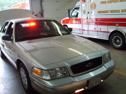 2004 Ford Crown Victoria - A silver police car with its hood open and emergency lights flashing sits in a garage next to an ambulance.
