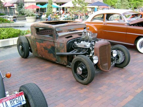 1974 Ford Ranchero - A chopped, heavily patinated rat rod hot rod coupe sits at a car show on a brick-paved plaza, showing rusty bare metal and black bodywork w…