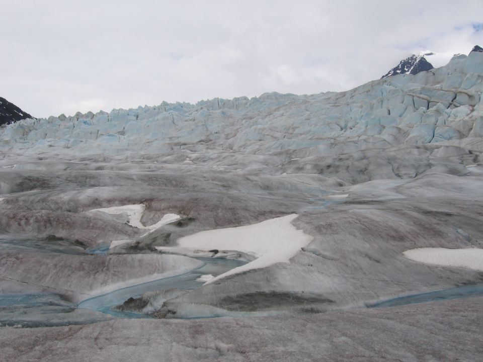 Expansive glacier surface with blue meltwater streams flowing through grey ice and snow, mountains in background