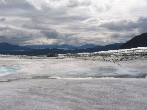 1973 Ford Ranchero - A vast glacial landscape unfolds with icy terrain, meltwater pools, scattered rocks, and distant mountains beneath a dramatic cloudy sky.