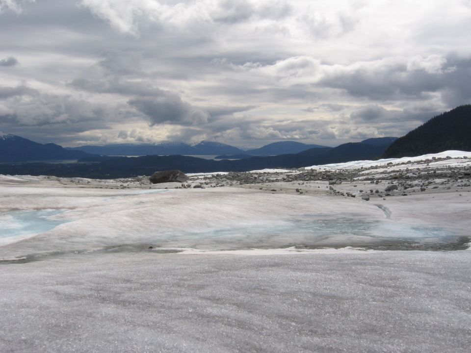 Glacial landscape with ice, meltwater pools, rocks, and distant mountains under a cloudy sky