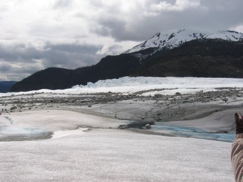 1973 Ford Ranchero - A vast glacier stretches towards snow-capped mountains under a cloudy sky, with a person's hand visible in the lower right corner.