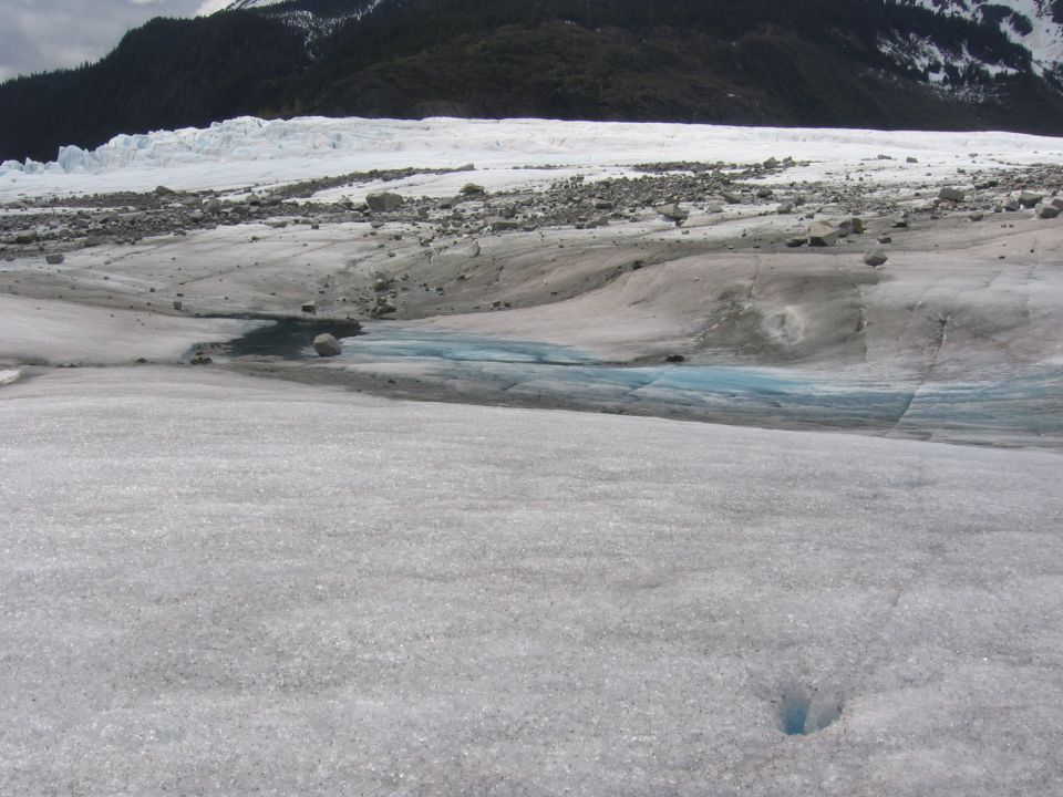 Glacier surface with blue meltwater pools and rocky debris, mountains in background