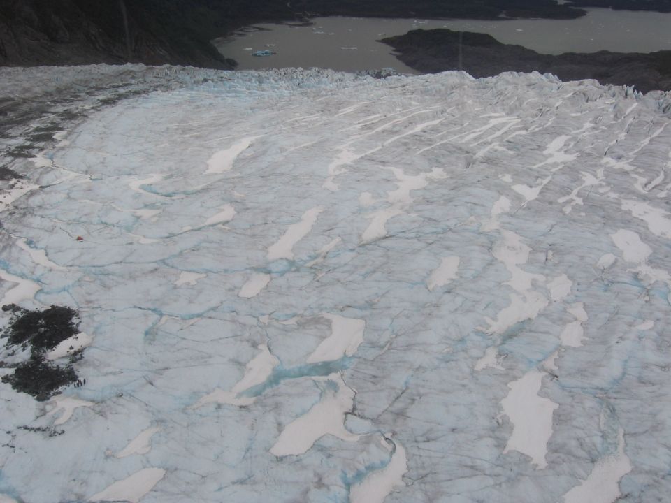 Glacier ice with blue crevasses and patches of snow, leading to a dark lake and forested hills