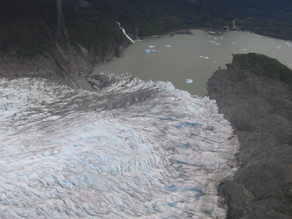 Aerial view of a large glacier flowing into a lake with icebergs. Mountains and forest are in the background