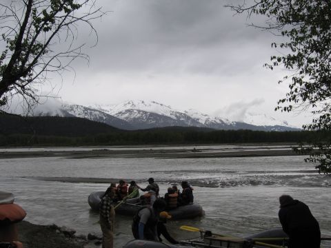 1973 Ford Ranchero - A group prepares for a rafting trip on a wide river, with a dramatic backdrop of snow-covered mountains under a cloudy sky.