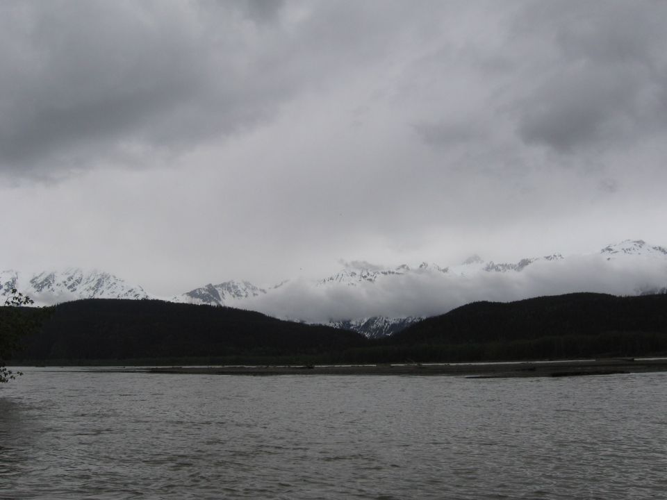 Overcast sky above snow-capped mountains and dark forested hills, with a wide river in the foreground