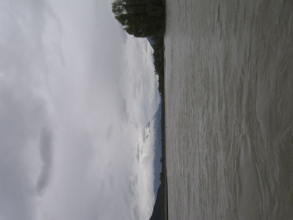 River flows past a tree line with snow-capped mountains under a cloudy sky