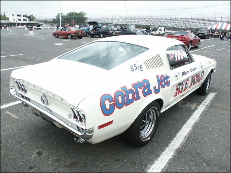 White 1967 Ford Mustang race car with 'Cobra Jet' and 'Rye Ford' graphics, rear view. Other cars and bleachers in background