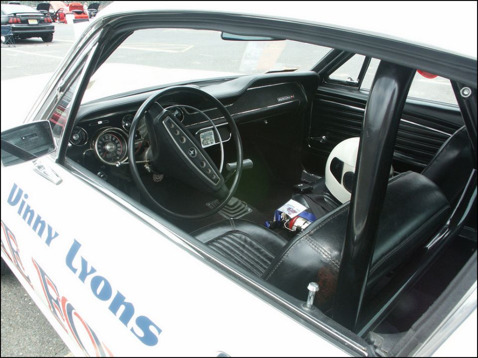 White car interior with black racing seats, roll cage, and a white helmet on the passenger seat. "Vinny Lyons" is written on the door