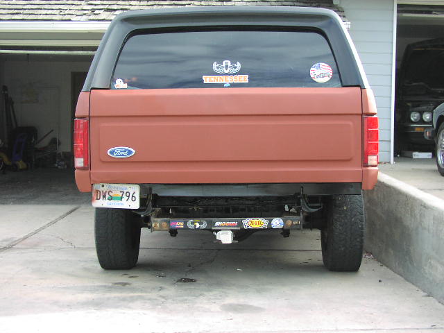 Rear of a primer-red Ford Bronco with black hardtop and sticker-covered rear bumper in a driveway