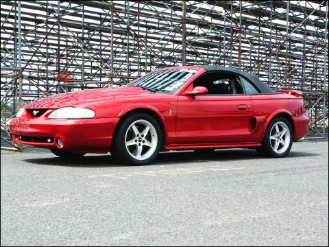 2001 Mercury Grand Marquis - A red convertible Mustang is parked on asphalt with stadium seating in the background.