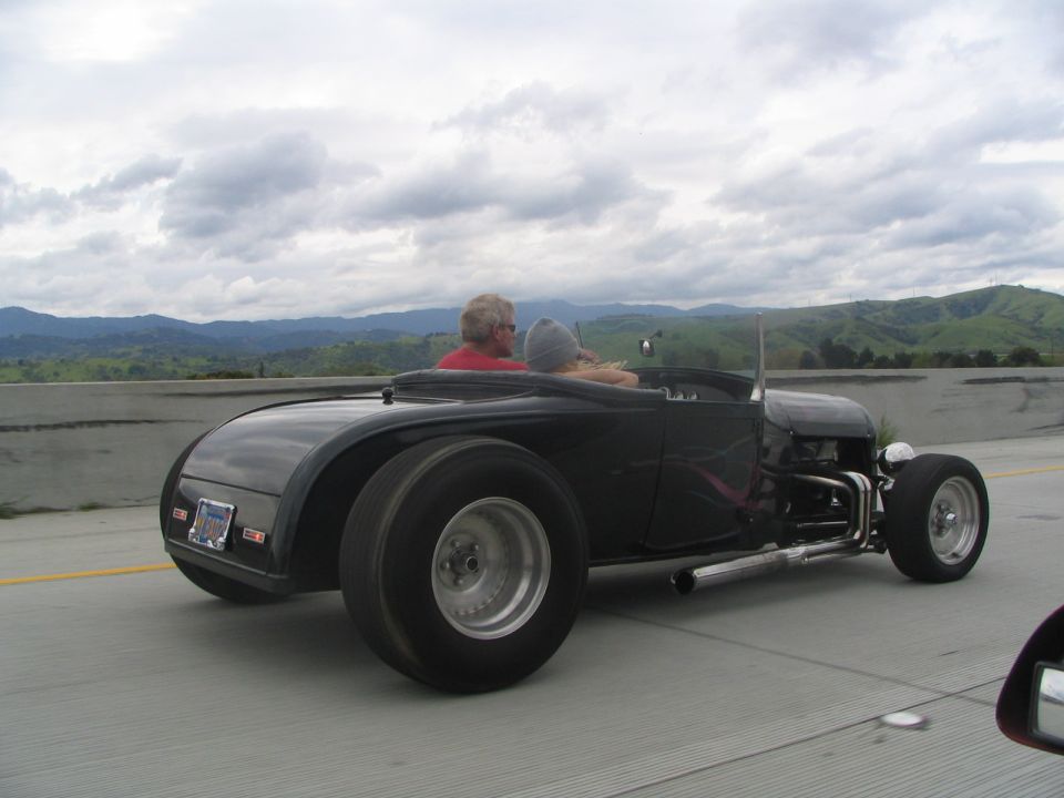 Black hot rod roadster with exposed engine and large rear tires, driving on highway