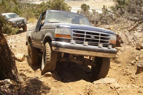 1986 Ford Bronco - A dark blue Ford Bronco tackles a challenging off-road path with another vehicle visible in the background.
