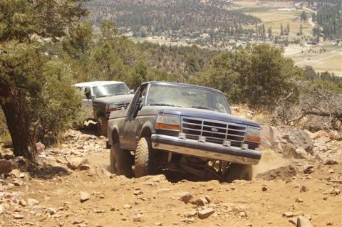 1986 Ford Bronco - Two Ford trucks navigate a challenging, rocky off-road path with a scenic valley in the background.