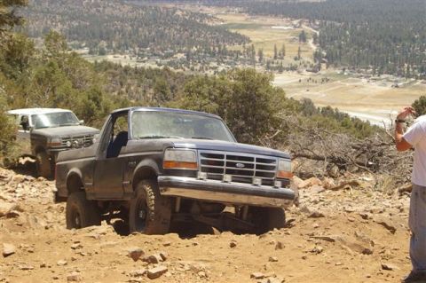 1986 Ford Bronco - Two Ford Broncos navigate a steep, rocky off-road trail with a scenic valley below.