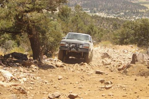 1986 Ford Bronco - A gray SUV navigates a challenging, rocky off-road trail with a scenic overlook in the background.